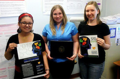 Elizabeth Sanchez, Tara Perkins, and Jamie Harguess posing in front of SRS poster while holding the symposium program.