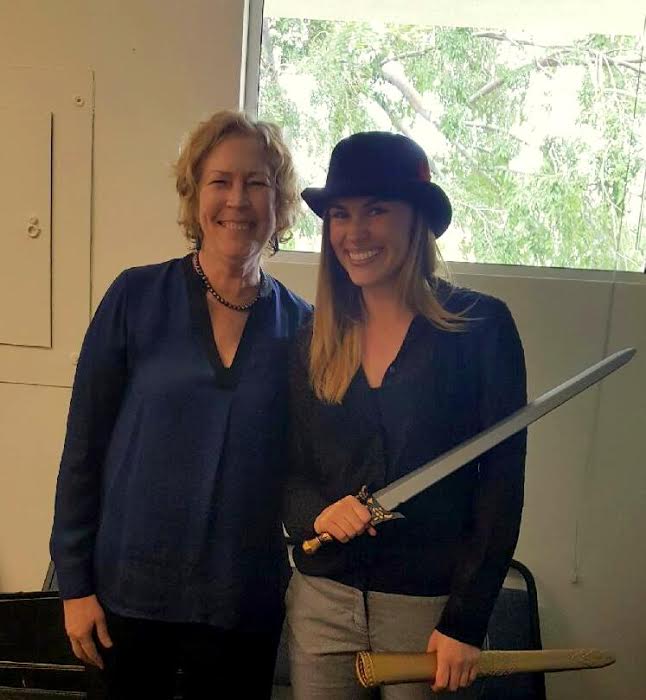 Smiling Maggie and mentee kristi who is holding sword in front of window